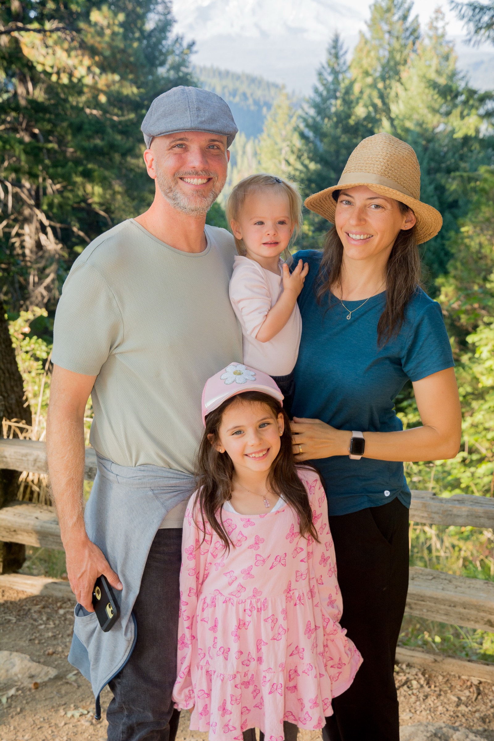 Joel, Maria, and their two daughters on a forest trail in front of a snow-capped mountain